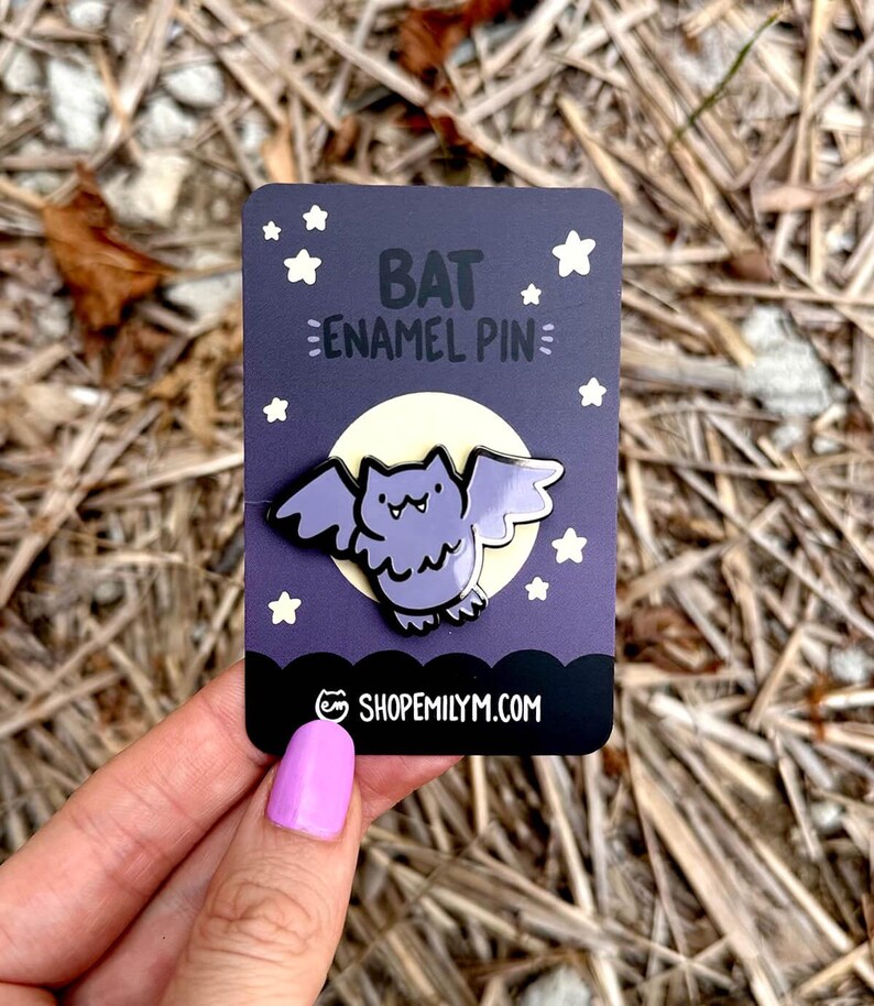 May include: Purple enamel pin of a cartoon bat with a yellow moon and white stars in the background. The text "BAT ENAMEL PIN" is written in black on the card. The card also has the website "SHOPEMILYMM.COM" written on it.