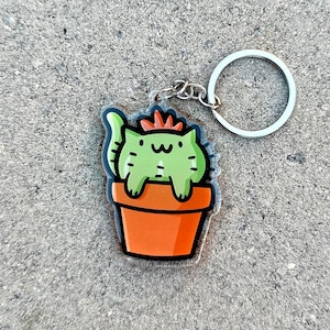 May include: A green cat-shaped cactus keychain with a red flower on top, sitting in an orange pot. The keychain is attached to a silver key ring.