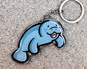 May include: A light blue manatee keychain with a black outline. The manatee has a smiling face with freckles and a pink tongue. The keychain includes a silver metal keyring and chain.