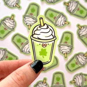 May include: A green and white cartoon shamrock shake with a straw and whipped cream. The shake has a happy face and a four-leaf clover on the front.