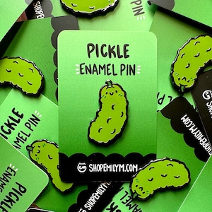 Puede incluir: Un pin de esmalte verde con forma de pepinillo con una cara sonriente. El pin dice "Pickle Enamel Pin" e incluye el sitio web "shopemilym.com".