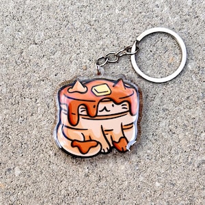 May include: A clear acrylic keychain featuring a cartoon cat wearing a pancake hat with a pat of butter on top. The cat is orange and white with a brown pancake hat and a yellow pat of butter.