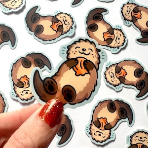 Op de afbeelding: Een verzameling stickers met een cartoon otter ontwerp. De stickers tonen otters in verschillende poses, met bruine vacht, lichtbruine buiken en het vasthouden van oranje objecten. Elke sticker heeft een lichtblauwe omtrek.