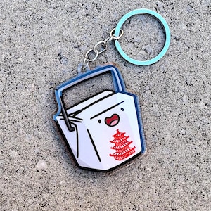 May include: A white and black takeout container keychain with a red pagoda design and a smiling face. The keychain has a silver chain and a light blue ring.