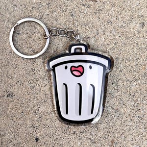 May include: A clear acrylic keychain featuring a cartoon trash can design. The trash can is white with a black outline, a smiling face, and a red mouth. The keychain includes a silver keyring and chain.