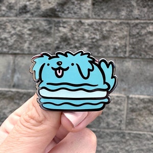 May include: A blue enamel pin of a cartoon dog wearing a blue and white macaron. The dog has a happy expression and is sticking its tongue out.