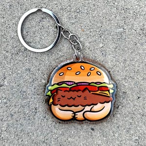 May include: A keychain featuring a cartoon cat wearing a hamburger costume. The cat is brown and has a white belly. The hamburger is yellow, brown, and green. The keychain is made of clear acrylic.