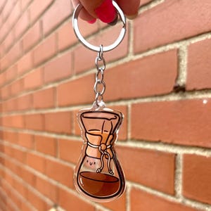 Op de afbeelding: Een transparante acryl sleutelhanger met een cartoonillustratie van een koffiefilter met een glimlach. Het koffiefilter is gevuld met bruine koffie en heeft een bruin papieren filter.