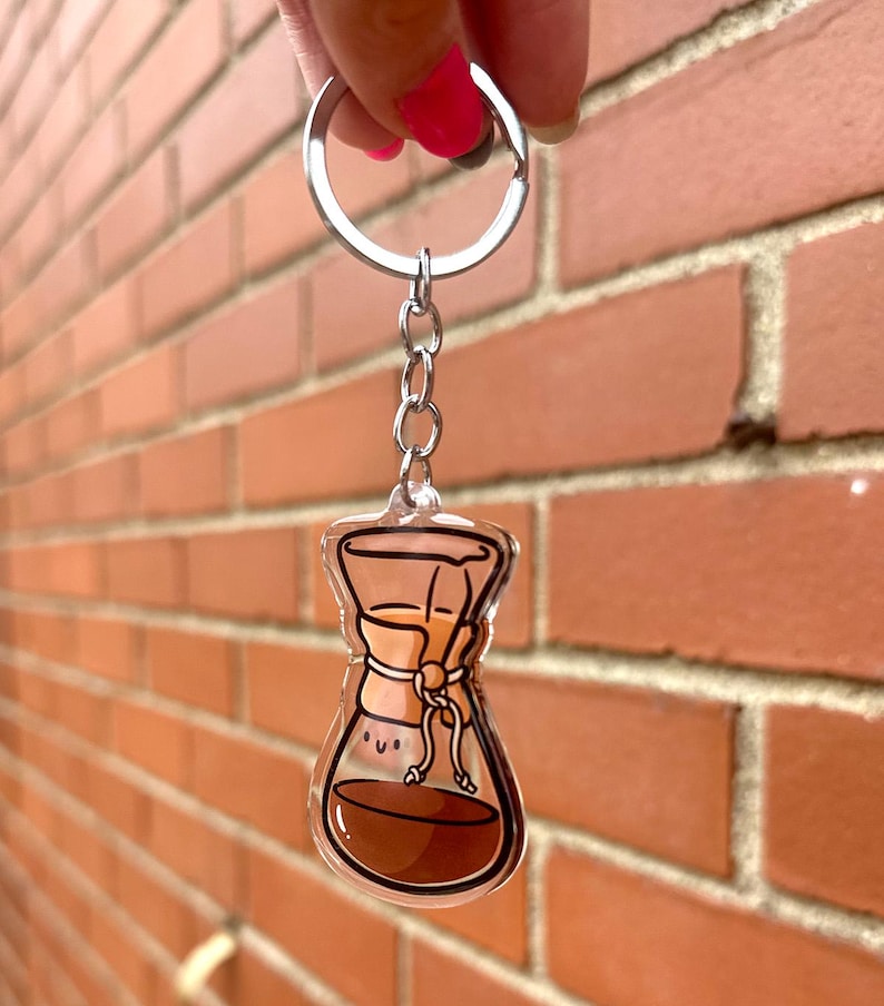 Op de afbeelding: Een transparante acryl sleutelhanger met een cartoonillustratie van een koffiefilter met een glimlach. Het koffiefilter is gevuld met bruine koffie en heeft een bruin papieren filter.