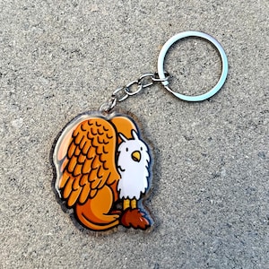 May include: A keychain featuring a cartoon griffin with orange and brown wings and a white body. The griffin is holding its wings behind its back and has a small, brown tail.