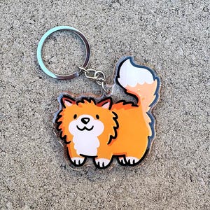 May include: A clear acrylic keychain featuring a cartoon illustration of a fluffy orange dog with a white chest and tail. The keychain has a silver metal ring and clasp.