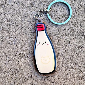 May include: A milk bottle keyring with a red cap, smiling face, and a cat face design. The keyring has a light blue ring and silver chain. The milk bottle is white with a black outline.