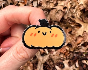 以下が含まれることがあります： 黒い縁取りのある、オレンジ色のカボチャのエナメルピン。カボチャは、頬が赤く、黒い茎のある笑顔の顔をしています。茶色の葉を背景にピンが配置されています。