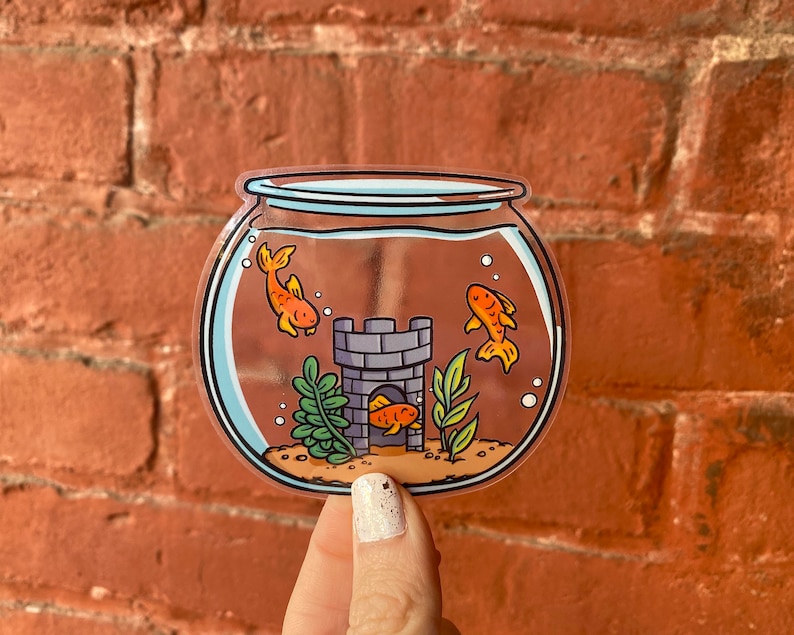May include: A clear glass fishbowl with three orange goldfish swimming inside. The fishbowl has a small grey castle at the bottom with a green plant.