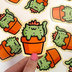 May include: A set of colorful stickers featuring cartoon cats in potted plants. The stickers are printed on white paper and have a glossy finish. The cats are green and have orange pots. The text on the stickers reads "Shop Emily".