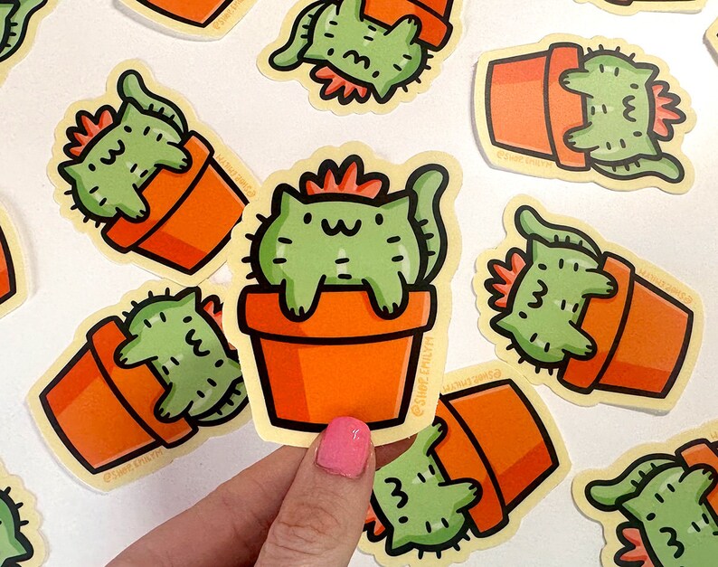 May include: A set of colorful stickers featuring cartoon cats in potted plants. The stickers are printed on white paper and have a glossy finish. The cats are green and have orange pots. The text on the stickers reads "Shop Emily".