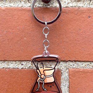 Op de afbeelding: Een bruine en transparante acryl sleutelhanger in de vorm van een koffiefilter met een glimlachend gezicht. De sleutelhanger is bevestigd aan een zilveren metalen ring en ketting.