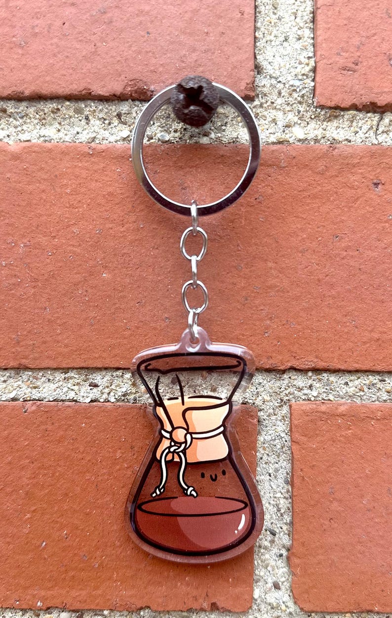 Op de afbeelding: Een bruine en transparante acryl sleutelhanger in de vorm van een koffiefilter met een glimlachend gezicht. De sleutelhanger is bevestigd aan een zilveren metalen ring en ketting.