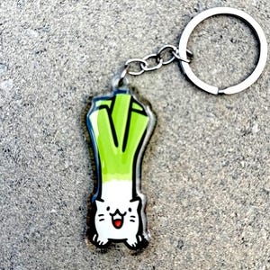 May include: A green and white keychain shaped like a leek with a cartoon cat face. The keychain has a silver metal ring.
