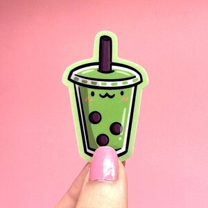 May include: Green and white cartoon bubble tea with black boba pearls and a cute face. The bubble tea is in a clear cup with a green lid and a straw.
