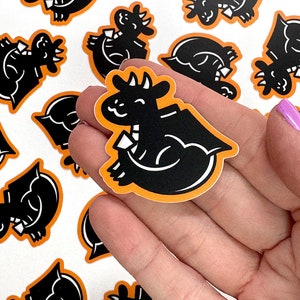 May include: A hand holding a black and orange sticker featuring a cartoon dragon design. The dragon is facing right and has a small wing on its back. The sticker is surrounded by other stickers with similar designs.