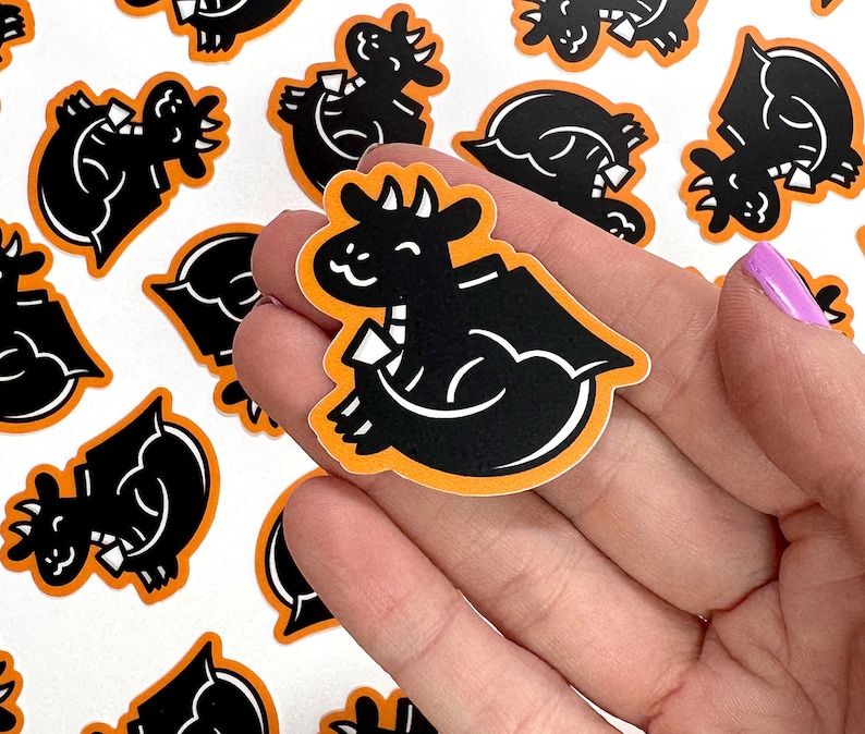 May include: A hand holding a black and orange sticker featuring a cartoon dragon design. The dragon is facing right and has a small wing on its back. The sticker is surrounded by other stickers with similar designs.