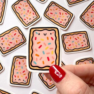 May include: A collection of rectangular stickers featuring a cartoon pop-tart design. Each sticker has a pink filling with colorful sprinkles, a brown crust, and a smiling face. The stickers are held by a hand with red nail polish.