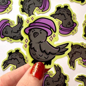 May include: A collection of stickers featuring a cartoon crow wearing a purple witch hat. The crow is gray with black outlines, set against a lime green background with diamond accents. The stickers are held by a hand with red nail polish.