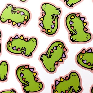 Op de afbeelding: Een set van 12 groene dinosaurus stickers met roze contouren en kleurrijke accenten. Elke dinosaurus heeft een glimlachend gezicht en een staart met een rij kleurrijke driehoeken.