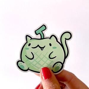 May include: A green and white cartoon cat sticker with a melon-like pattern and a small green stem on its head.
