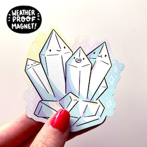 May include: A weatherproof magnet featuring a cluster of white crystal shapes with cute cartoon faces. The crystals are outlined in pastel colors and have a sparkly background.