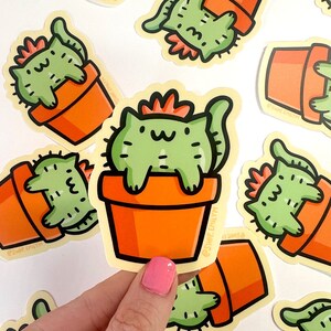 May include: A set of stickers featuring a cartoon cat dressed as a cactus in a terracotta pot. The cat is green with a red crown and a white face. The stickers are on a white background and have the text "@shop.earthly" on them.