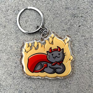 Puede incluir: Un gato negro con cuernos de diablo rojos y una capa roja está rodeado de llamas amarillas. El gato lleva un pañuelo rojo y tiene una expresión traviesa. La imagen es un llavero.
