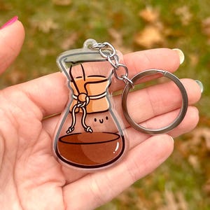 Op de afbeelding: Een transparante acryl sleutelhanger in de vorm van een koffiefilter met bruine koffie erin en een schattig gezicht. De sleutelhanger heeft een zilveren metalen ring.