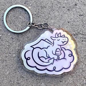 May include: A white dragon with a pink cloud and stars, printed on a clear acrylic keychain. The keychain has a silver metal ring and chain.