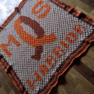 May include: A crocheted blanket with an orange ribbon and the words "My Warriors" in brown and orange yarn.