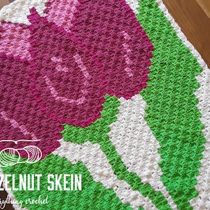 May include: A crochet blanket featuring a design of three pink tulips with green stems and leaves on a white background. The blanket is made with a textured stitch and has a scalloped edge. The text "THE HAZELNUT SKEIN" is visible in the bottom left corner of the image.