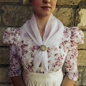 May include: A white blouse featuring a purple floral pattern, puff sleeves, and a sheer white neck scarf secured with a round, gold-toned filigree brooch. The garment is worn with a plain white skirt, creating a layered look with distinct textures.