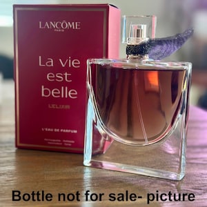 Puede incluir: Un frasco de perfume de vidrio transparente con un líquido ámbar oscuro, una caja cuadrada y una parte superior decorativa. La caja es de color rojo intenso con las palabras "Lancôme Paris" y "La vie est belle" impresas en blanco. El frasco tiene una tapa transparente y un acento decorativo púrpura oscuro.
