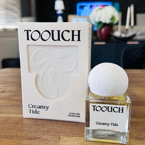 May include: A rectangular white perfume box and a square glass perfume bottle with a white textured spherical cap. The box and bottle label both feature the black text TOOUCH and Creamy Tide, with the box also displaying EAU DE PARFUM.