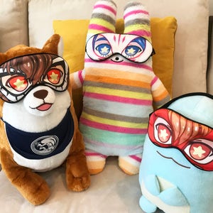 May include: Three plush toys wearing sleep masks with colorful star designs. The toys are a brown dog, a striped bunny, and a blue egg-shaped character.