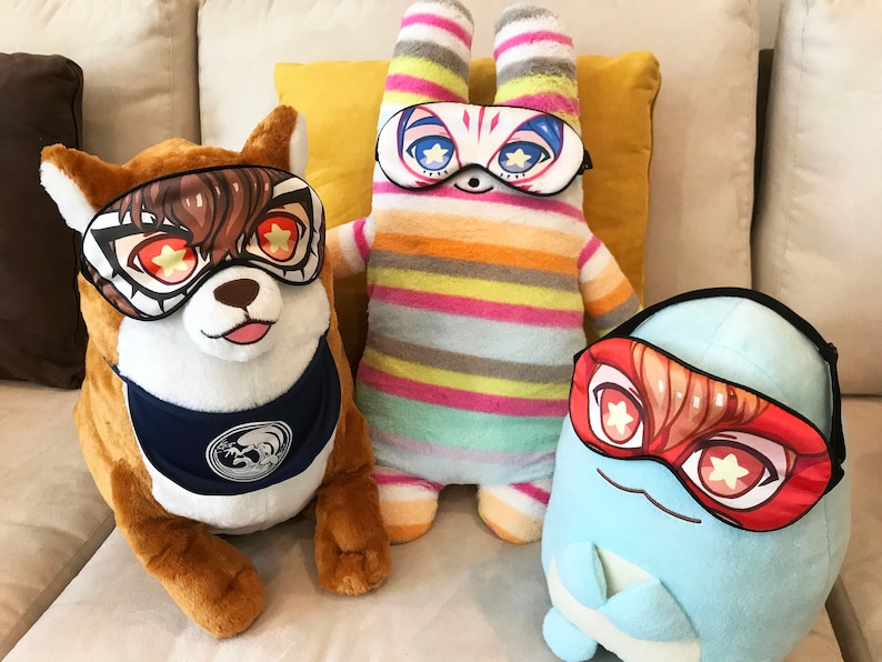 May include: Three plush toys wearing sleep masks with colorful star designs. The toys are a brown dog, a striped bunny, and a blue egg-shaped character.