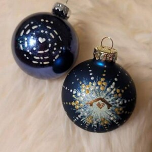 May include: Two blue glass Christmas ornaments with white and gold painted designs. One ornament has a white heart and a swirling pattern. The other ornament has a white and gold starburst pattern with a small house in the center.