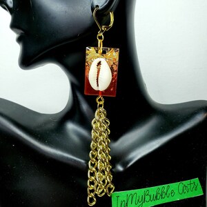 May include: Gold-toned dangle earrings with a rectangular red and gold resin pendant. Each earring features a white seashell and a cascade of gold chain tassels. The earrings are displayed on a black mannequin head.
