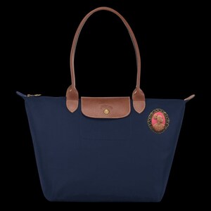 May include: A navy blue tote bag with brown leather handles and a gold zipper. The bag has a brown leather patch with a gold logo on the front and a pink cameo with a gold frame.