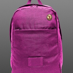 May include: A magenta backpack with a small, round, gold-colored pin on the front. The backpack has a zippered pocket on the front and a large main compartment.