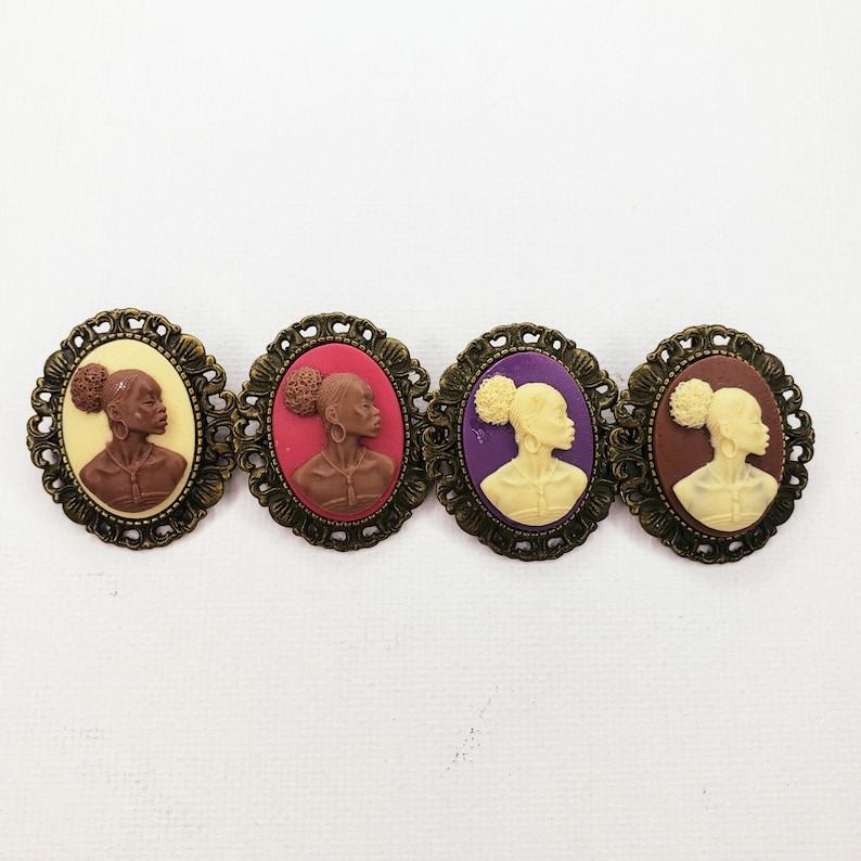 May include: Four cameo brooches with gold-tone frames. Each brooch features a different colored background: pink, purple, brown, and cream. Each cameo features a profile of a woman with a bun hairstyle.