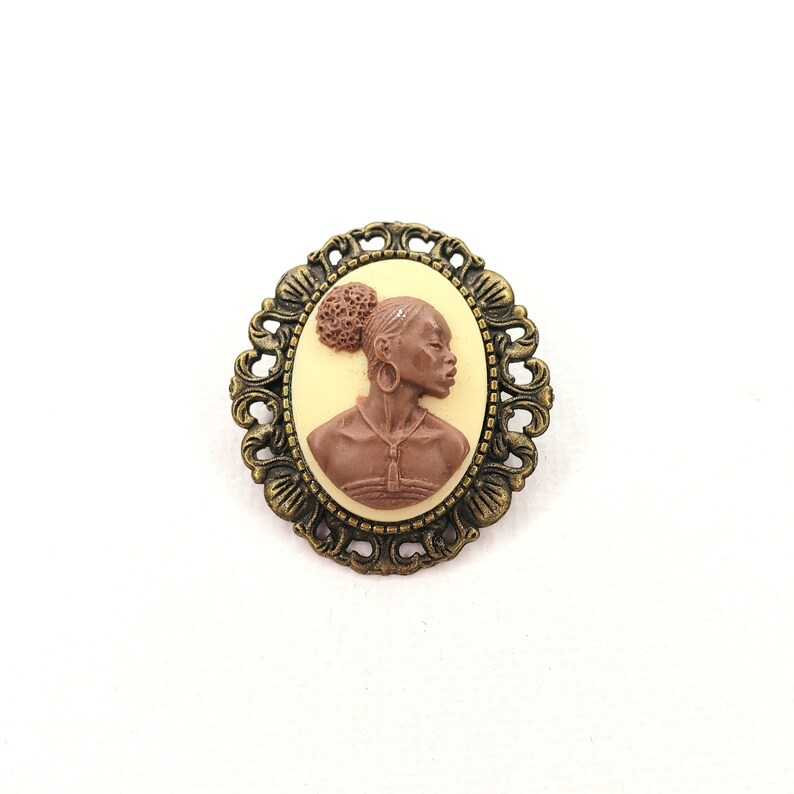 May include: A brown cameo brooch with a detailed profile of a woman with a bun in her hair. The cameo is set in a brass-toned ornate metal frame.