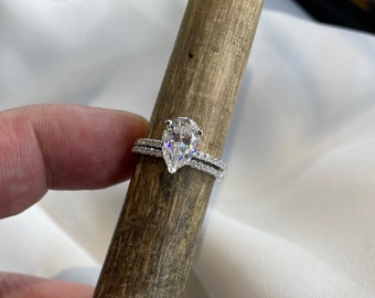 May include: A pear-shaped diamond engagement ring with a silver band and a matching wedding band. The ring is set on a wooden surface.