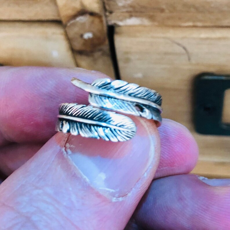 Puede incluir: Anillo de plata con dos plumas envueltas una alrededor de la otra. El anillo es ajustable y se puede llevar en cualquier dedo.